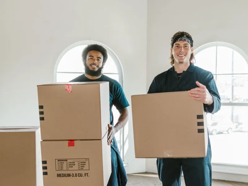 Two removal men in navy uniforms carrying cardboard boxes through an empty house